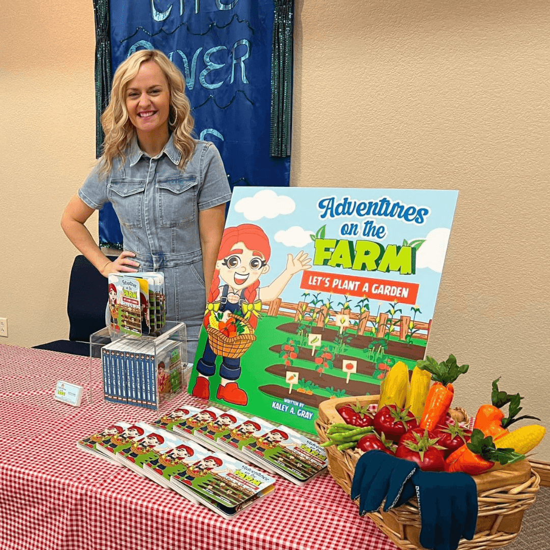 Author at book signing table for 'Adventures on the Farm: Let's Plant a Garden' children's book, featuring colorful farm-themed display and fresh vegetables.